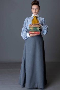 Çizgili pamuklu bluz, hardal sarısı kravat ve gri etek giyen Edwardlı işçi sınıfı bir kadın elinde kitaplarla stüdyo arkaplanına yaslanıyor.