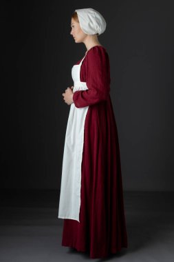 Kırmızı keten elbise giyen, önlüklü ve şapkalı bir hizmetli stüdyo arkaplanına yaslanmış.