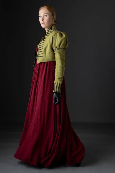 スタジオを背景に緑の編組のスペンサーと手袋をした赤いリネンのドレスを着たリージェンシー女性 — ストック写真
