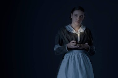 Bir hizmetçi ya da işçi sınıfı Viktorya dönemi kadını koyu yeşil giyinmiş korse ve etek giyiyor ve elinde mum tutuyor.