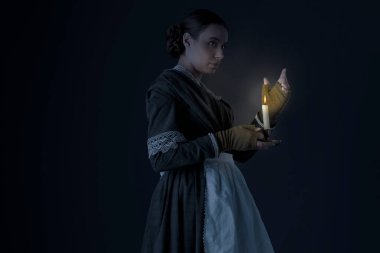 Bir hizmetçi ya da işçi sınıfı Viktorya dönemi kadını koyu yeşil giyinmiş korse ve etek giyiyor ve elinde mum tutuyor.