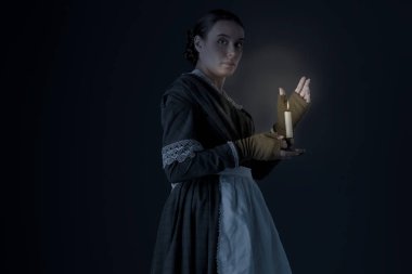 Bir hizmetçi ya da işçi sınıfı Viktorya dönemi kadını koyu yeşil giyinmiş korse ve etek giyiyor ve elinde mum tutuyor.