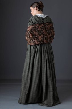 Koyu yeşil korse ve şal desenli etek giyen Viktorya dönemi işçi sınıfı bir kadın.