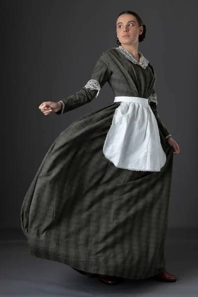 一位维多利亚时代的工人阶级妇女 身穿深绿色的格子裙和带花边领子的裙子 围裙衬托着灰色的工作室背景 — 图库照片
