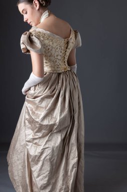 Uzun, dantel eldivenli altın renkli balo elbisesi giyen ve gri bir fonda duran Viktorya dönemi kadını.