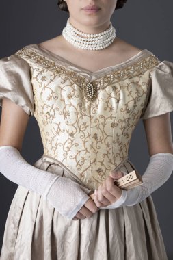 Uzun, dantel eldivenli altın renkli balo elbisesi giyen ve gri bir fonda duran Viktorya dönemi kadını.