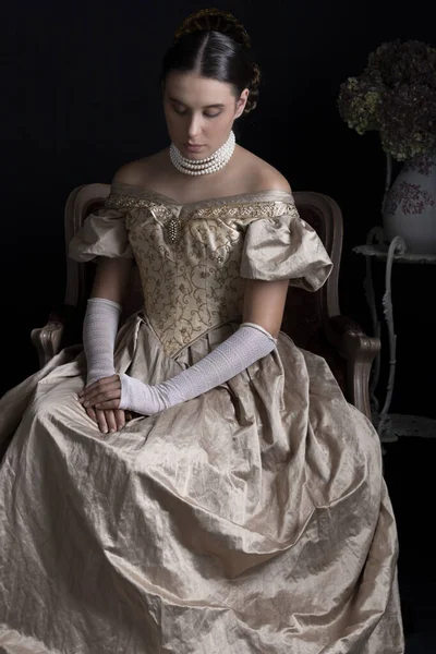 Altın renkli balo elbisesi giyen ve antika bir sandalyede oturan Victoria döneminden kalma bir kadın.