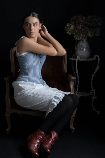 一个维多利亚时代的女人 穿着紧身胸衣和各种衣服坐在昏暗的房间里的椅子上 — 图库照片