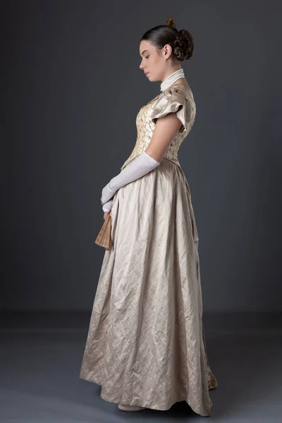 A Victorian woman wearing a gold ball gown with long, lace fingerless gloves and standing against a grey backdrop