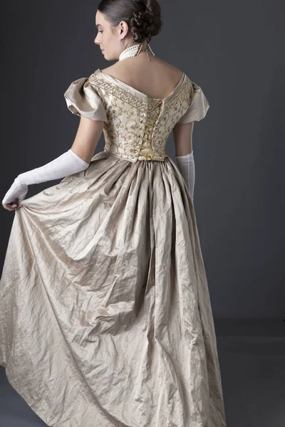Uzun, dantel eldivenli altın renkli balo elbisesi giyen ve gri bir fonda duran Viktorya dönemi kadını.
