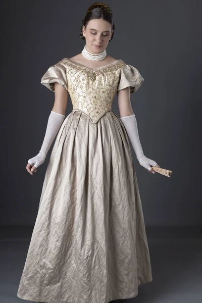 A Victorian woman wearing a gold ball gown with long, lace fingerless gloves and standing against a grey backdrop