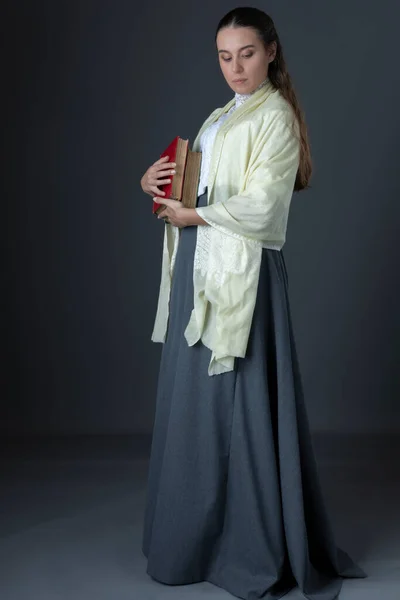 A Victorian or Edwardian woman standing and holding a pile of books against a studio backdrop