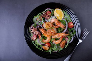 Deniz ürünleri, roka, domates, salatalık, kırmızı soğan ve limonlu siyah tabak. Siyah arkaplan, yakın plan.