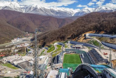 Soçi, Rusya - 1 Nisan 2014: Ruski Gorki Kayak Merkezi 'nin ilkbaharda dağ ormanı ve karlı zirvelerle çevrili güneşli manzaralı manzarası, kayak atlayışlarının tepesinden dikey vew.