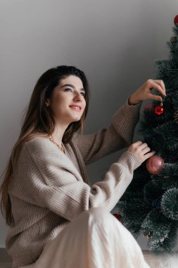 Kahverengi kazak giyen genç güzel esmer Noel ağacını oyuncaklarla süslüyor ve bayram beklentisiyle gülümsüyor ve çok mutlu. Mutlu yıllar, tatil geçmişi