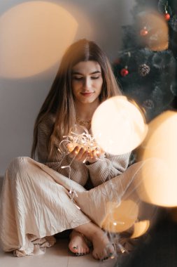 Güzel esmer, bej bir kazak giymiş elinde altın bir çelenkle poz veriyor ve ön planda büyük bir bokeh var. O, çelenge bakıyor ve mutlu. Yeni yıl ve Noel konsepti