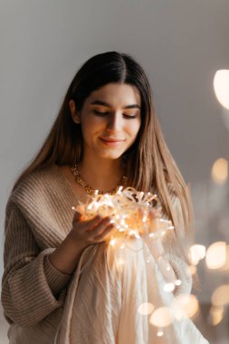 Güzel esmer, bej bir kazak giymiş elinde altın bir çelenkle poz veriyor ve ön planda büyük bir bokeh var. O, çelenge bakıyor ve mutlu. Yeni yıl ve Noel konsepti