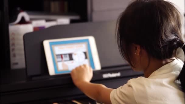 小アジアの女の子は ピアノで練習しながら アプリケーションとビデオストリーミングのオンラインピアノ音楽レッスンを探しています — ストック動画