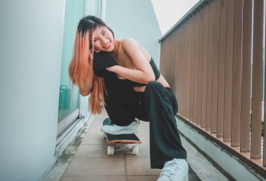 Asyalı patenci manken kız beyaz spor ayakkabılar ve kaykayla havalı bir sokak sporu modası için balkonda poz veriyor.