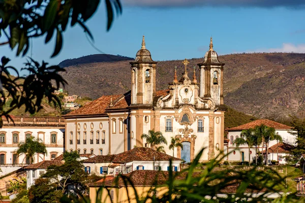Ouro Preto şehrinin en büyük tarihi kiliselerinden biri arka planda dağlarla birlikte bitki örtüsünün içinden görülüyor.