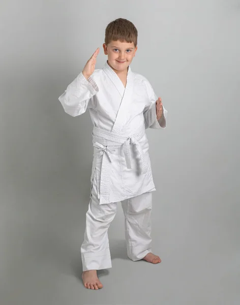 Niño en kimono blanco sonríe y se para en la postura defensiva de combate contra el fondo gris —  Fotos de Stock