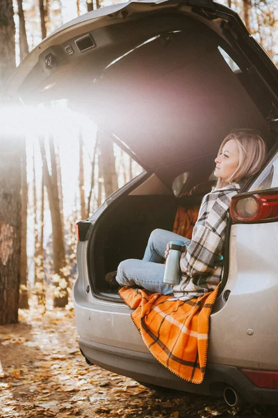 a blonde adult girl in a plaid shirt is drinking coffee sitting in the open trunk of a car. picnic in the car.