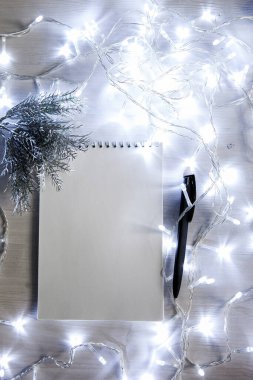 Noel çelengi içinde gümüş renkli bir köknar ağacı dalının olduğu bir defter, siyah bir kalem. ışık arkaplanı