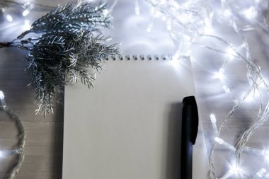 Noel çelengi içinde gümüş renkli bir köknar ağacı dalının olduğu bir defter, siyah bir kalem. ışık arkaplanı