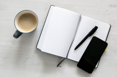 Beyaz bir defter, siyah bir cep telefonu ve açık ahşap arka planda bir fincan kahve.