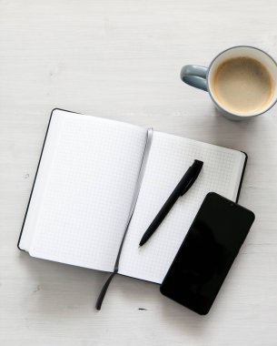 Beyaz bir defter, siyah bir cep telefonu ve açık ahşap arka planda bir fincan kahve.