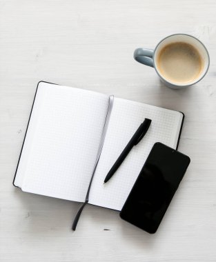 Beyaz bir defter, siyah bir cep telefonu ve açık ahşap arka planda bir fincan kahve.