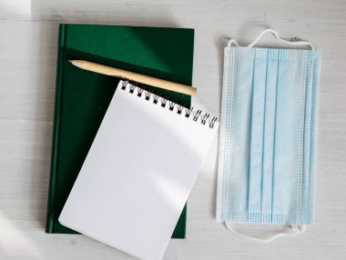 Yeşil bir günlükte açık beyaz bir defter, arka planda çevre dostu bir kalem, tıbbi bir maske ve yeşillik dalı.