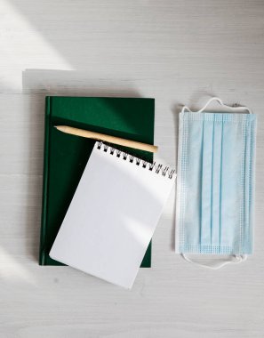 Yeşil bir günlükte açık beyaz bir defter, arka planda çevre dostu bir kalem, tıbbi bir maske ve yeşillik dalı.