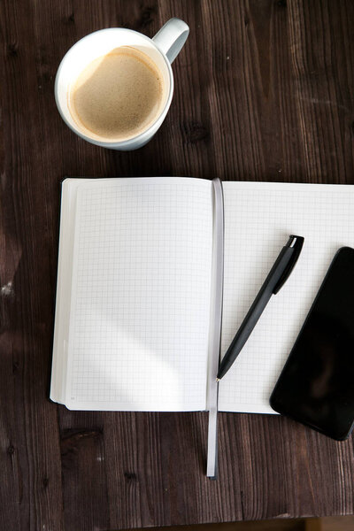 open white notebook , a black mobile phone and a cup of coffee on a dark wooden background
