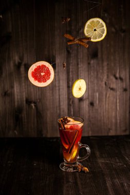 Noel şarabı, şeffaf bir bardakta, koyu ahşap bir zemin üzerinde, uçan baharatlar ve meyvelerle çevrili.