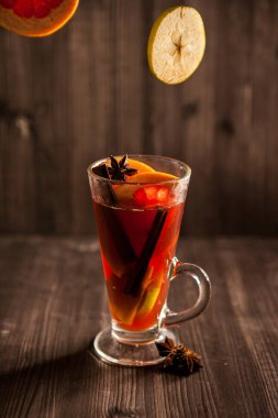 Noel şarabı, şeffaf bir bardakta, koyu ahşap bir zemin üzerinde, uçan baharatlar ve meyvelerle çevrili.