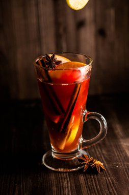 Noel şarabı, şeffaf bir bardakta, koyu ahşap bir zemin üzerinde, uçan baharatlar ve meyvelerle çevrili.