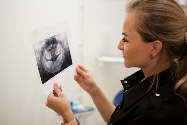 Siyah üniformalı, uzun sarı saçlı dişçi kadın elinde çenesinin röntgenini tutuyor.