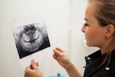 Siyah üniformalı, uzun sarı saçlı dişçi kadın elinde çenesinin röntgenini tutuyor.