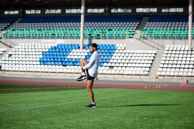 Siyah şortlu beyaz adam ve beyaz tişört, stadyumun yeşil çimlerinde koşmadan önce esniyor.
