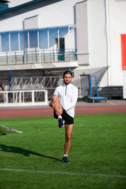 Siyah şortlu beyaz adam ve beyaz tişört, stadyumun yeşil çimlerinde koşmadan önce esniyor.