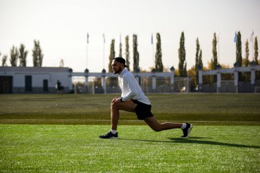 Siyah şortlu beyaz adam ve beyaz tişört, stadyumun yeşil çimlerinde koşmadan önce esniyor.