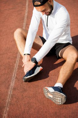 Beyaz elbiseli erkek koşucu dizini stadyumda tutuyor, spor sakatlığı var.