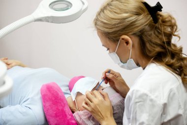 Bir kız, salgın sırasında kirpiklerini uzatma prosedürünü uygular. Elinde aletler olan bir usta.