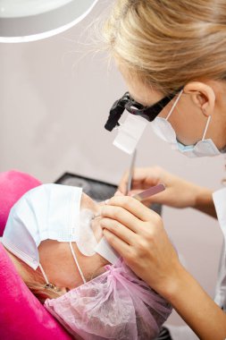 Bir kız, salgın sırasında kirpiklerini uzatma prosedürünü uygular. Elinde aletler olan bir usta.