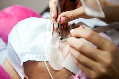 Bir kız, salgın sırasında kirpiklerini uzatma prosedürünü uygular. Elinde aletler olan bir usta.