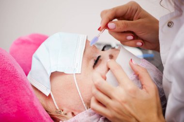 Bir kız, salgın sırasında kirpiklerini uzatma prosedürünü uygular. Elinde aletler olan bir usta.