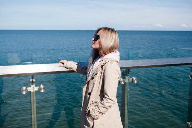 Soğuk bir denizin arka planında sırt çantalı sarışın bir kız, gökyüzünde martılar. seyahat ve seyahat konsepti, göçmenlik. Moda detaylarını kapat, plajda doluşan kız, günlük kıyafet, sırt çantası, gezgin, soğuk hava..