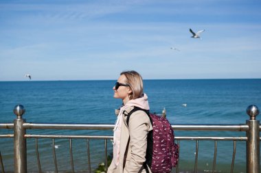 Soğuk bir denizin arka planında sırt çantalı sarışın bir kız, gökyüzünde martılar. Seyahat, seyahat, göç kavramı.