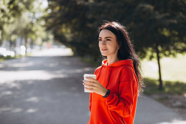 Stylish girl in red sweater drinking coffee in the city. Happy woman is waiting friends and relaxing in the park, outdoors, summer time